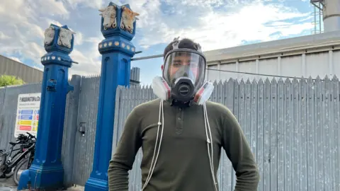 Aisha Iqbal/BBC A young man stands in front of a factory plant, identified by a sign in the background as P Waddington and Co. The lad wears a gas mask.