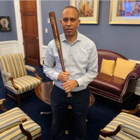 Hakeem Jeffries/Instagram US House Minority Leader Hakeem Jeffries holds a brown baseball bat while standing in an office. He is surrounded by chairs including a brown couch with yellow cushions which is behind him. He is wearing a blue dress shirt and black trousers