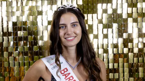 Head and shoulders image of Melisa Raouf wearing a tiara and sash that has the start of the word 'bare' on it. She is smiling and wearing no make up. She is standing in front of a gold, shimmering background