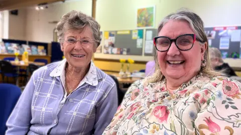 Muriel and Pamela women sit together indoors at a community space, smiling as they talk, both wear glasses and have brightly coloured tops.