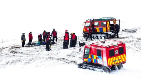 Braemar Mountain Rescue Team Red vehicles with tank-style caterpillar tracks, red body with white writing which says Braemar Mountain Rescue, and the word ambulance in red on blue and yellow checks, on snow, on a snowy hillside, with rescuers in bright mountain gear.