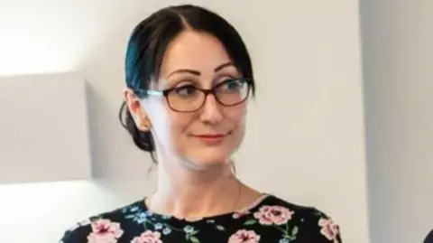 A woman with dark hair tied back, a split fringe and glasses is wearing a dark top with pink flowers on it. She is looking to her left.