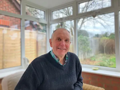 A man sitting in an outside conservatory and looking directly at the camera. He is relaxed and wearing a blue v-neck jumper and green shirt. There is a garden behind him outside to his left and he looks amiable and is smiling with a rosy complexion. 