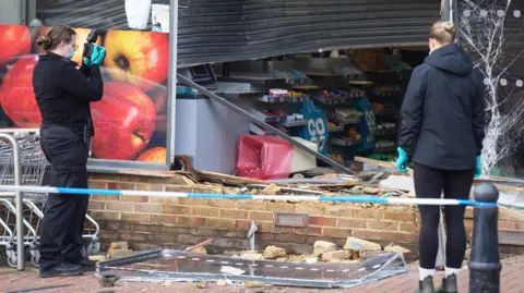Eddie Mitchell Two forensic officers taking photos and inspecting the outside of a damaged supermarket shop front. 