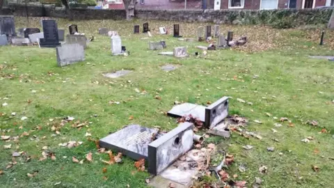 St George's Church, Stalybridge An open grassy cemetery with numerous headstones, some standing and others lying flat. In the foreground, two large gravestones are broken and toppled over, exposing their bases. Fallen leaves are scattered across the grass. A stone wall and residential houses can be seen in the background beyond the cemetery.