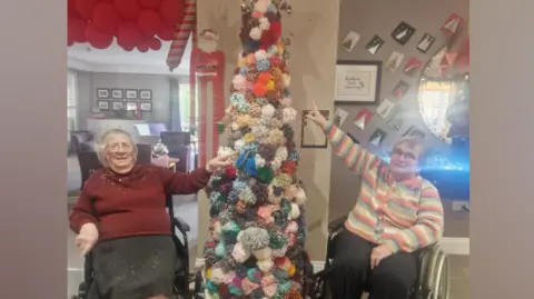 Vera Todd and Colette Roscamp are standing on each side of the colourful pom-pom Christmas tree with big star at its top. They are pointing at pom-poms they have made. Vera is wearing a red cardigan and a grey skirt and has shoulder-length white hair. Colette is wearing black trousers and colourful striped cardigan. She has short grey hair. Both are wearing glasses. The rest of the room is decorated for Christmas with lights and red stars hanging from the ceiling. 