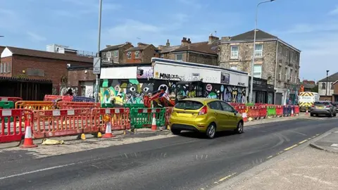BBC View of the roadworks on Park Street. There are multi-coloured crash barriers along one side of the road with cars waiting and a Northern Powergrid van parked on the corner of the junction