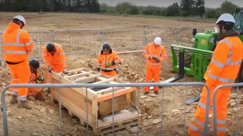 National Highways A stone coffin in a wooden frame next to a forklift truck with archaeologists in hi-vis orange jackets and trousers surrounding it