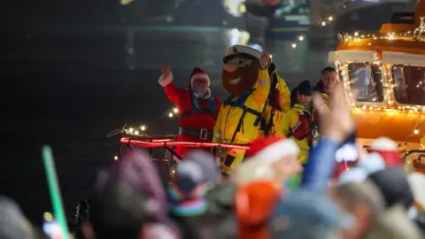 Penzance Council Father Christmas in his traditional red outfit stands next to a man wearing a lifeguard mascot costume, with yellow waterproofs and an oversized head with a brown beard on a lifeboat at night, while crowds of people wave from the shore.