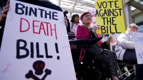 A group of anti-assisted dying protesters with signs, including one in the foreground that says "bin the death bill", with a skull and crossbones. 