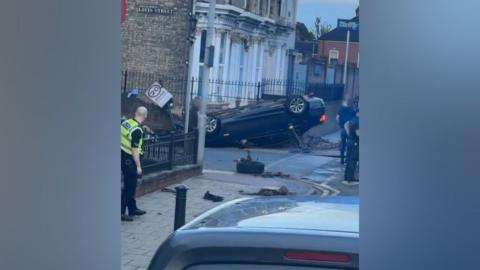 An overturned car next to a set of flats which has knocked down bollards and 20 zone sign with a policeman stood at the side
