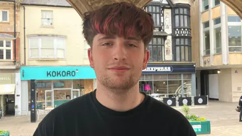 Joanna Taylor/BBC William who has dark brown hair and a longish red dyed fringe over his forehead. He is wearing a black T-shirt. He is standing in the Guildhall in Peterborough and through its arches is a row of shops with two rows of windows above. 