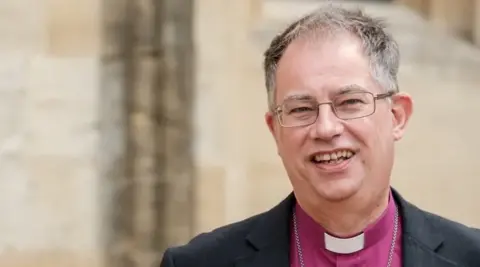 A man with grey hair and glasses is pictured wearing a clerical collar and smiling at the camera. 