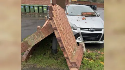 Matt Morris A brick wall  which has fallen over. Brick debris is on the windscreen of a silver Ford car.