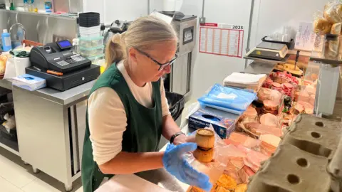 A woman wearing a green apron is preparing pork pies for a customer from behind a counter.