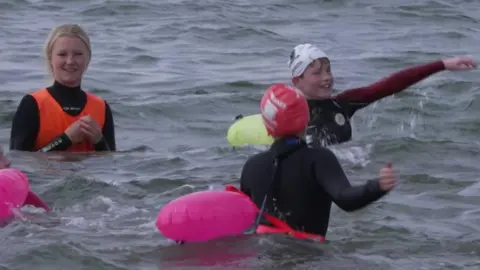 Three children in wetsuits with pink floats, splashing and smiling in the waves at the beach