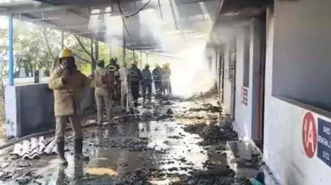 A first responder in a khaki uniform and yellow helmet stands inside a lobby where a blast has occurred. Other officials gather in the background and debris lies scattered across the floor.