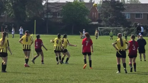 City of Gloucester WFC A girls' football game is taking place. One team are in yellow shirts, another is in red. The yellow football is high in the air after a kick. Behind is the goal, and behind that are houses behind the field.
