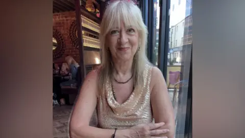 A woman with blonde hair and a fringe with pink-rimmed glassed on her head is smiling from someone across a table in what looks like a restaurant in the background. She is wearing a glittery beige and white top with a brown necklace.