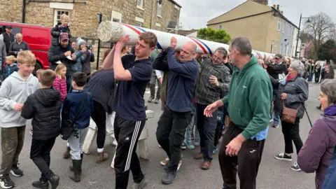 A large maypole being carried through a village by a group of men, as children play nearby.
