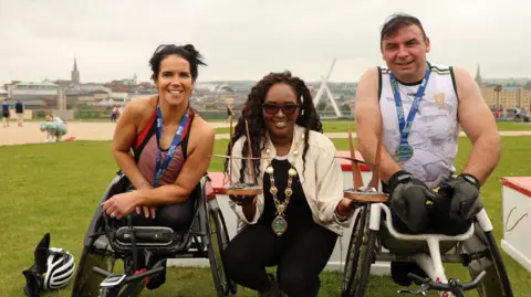 Derry City and Strabane District Council Lillian Seenoi Barr smiling between Jayne Bleakley who won the women’s wheelchair title and Karol Doherty who won the men’s wheelchair crown. Jayne and Karol are wearing their medals and smiling at the camera.