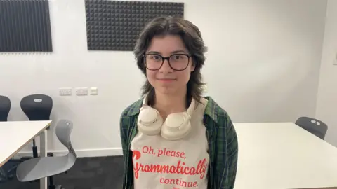 A young person who has shoulder length brown hair, and is wearing a white t-shirt with orange writing that reads "oh please, grammatically continue". They are also wearing a green plaid shirt and have black glasses and white headphones around their neck.