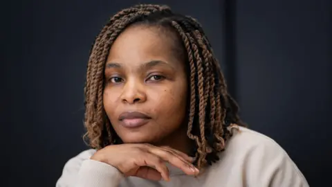 PA Media Ruvimbo Kavia – A woman of about 40 with braided hair and a cream top. She is looking towards the camera with her hand under her chin.