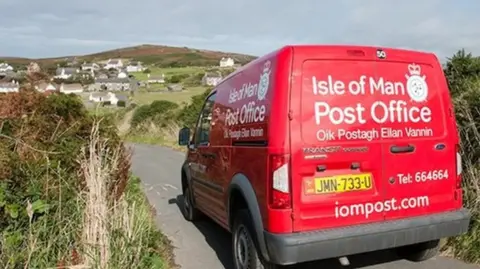 The back of a red postal van travelling on a country lane. A small cluster of white houses can be seen in the distance.