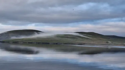 Kimberley Fairley A low hill covered with green grass has low lying grey clouds rolling over the top. There is a calm and still body of water in the foreground of the picture which reflects the blue and grey sky.