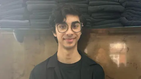 Adam Hussain painted in front of a shelf of folded black hair towels. He wears a black shirt over a black T-shirt and wears big black glasses. 