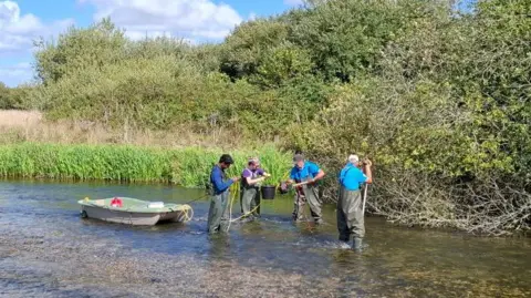 GWCT Four men in waders hold electro-fishing equipment and nets in the River Frome on a sunny summer day.