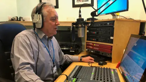 East Suffolk and North Essex NHS Foundation Trust John Alborough sits in a radio studio with headphones on. He is looking at a screen as he speaks into a microphone in front of him. He has grey hair and wears a blue shirt. One of his hands rests on the desk in front of him. 