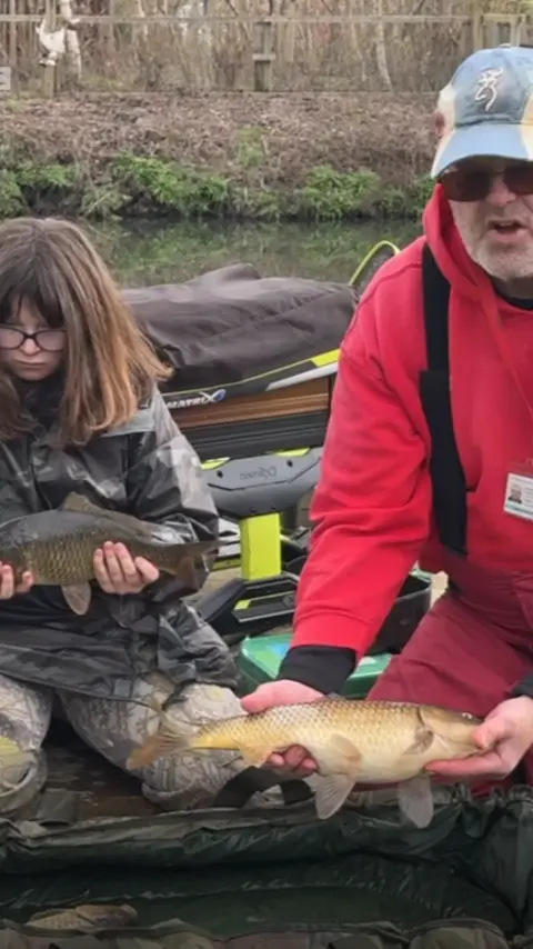 A man on the right wearing sunglasses, a grey cap and red hoodie, holding a fish out in front of him. On the left there is a girl with brown hair and glasses, wearing camouflage overalls and holding a smaller fish.