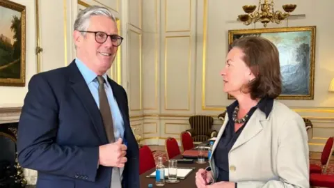 Sir Keir Starmer and Eluned Morgan stood in a meeting room with a chandelier, red chairs, a table and paintings hung on a wall.