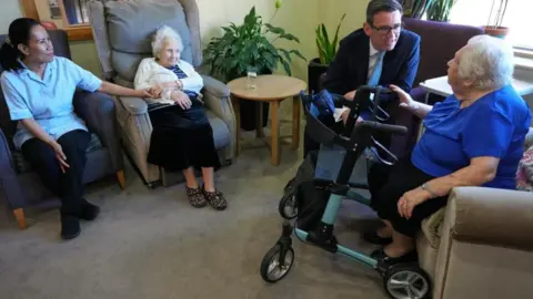 PA Media Andy Burnham is pictured sitting in a dark blue suit speaking to an elderly woman at a care home in north London. Another resident and a carer are pictured looking on.