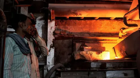 Getty Images Workers supervise a gas-run furnace inside a glass factory in India.