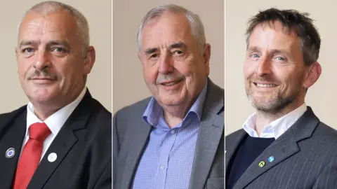 A Labour, SNP and Democratic Alliance councillor on Dumfries and Galloway Council. Labour's Archie Dryburgh on the left has a red tie and dark jacket, grey short hair and a moustache. On the right it the SNP's Stephen Thompson with a beard and spiky hair and an SNP badge on his jacket. In the centre is Willie Scobie of the Democratic Alliance in a blue shirt and grey jacket with grey hair.