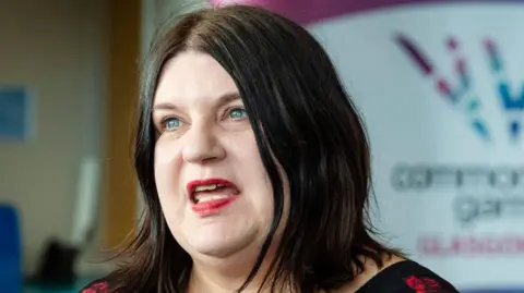 Susan Aitken has shoulder-length dark brown hair. She has blue eyes and is mid-speech