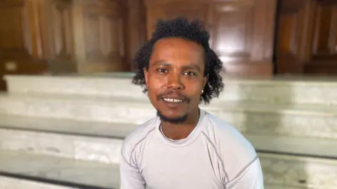 A man with black curly hair. He is wearing a white top. 