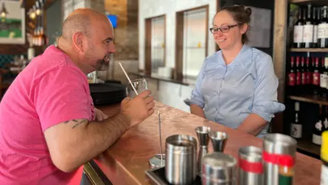 James stands at the bar with a glass in his hand with a white straw coming out of it. Amy is behind the bad and they're both laughing. Slightly blured in the foreground are some drinks shakers and measurers.