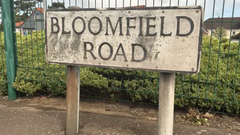 A road sign which reads "Bloomfield Road". It is on a footpath that has a green railing and grass behind it. 
