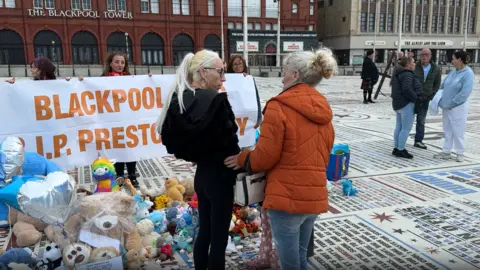 A middle aged woman with blonde hair tied back into a pony-tail stands alongside an older woman in an orange coat in front of a collection of teddy-bears and balloons.
