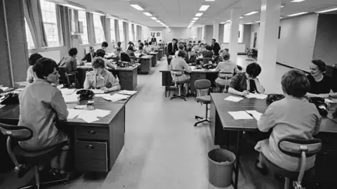 Getty Images A black and white photo inside the Barclaycard offices in the 1960s. There are rows of large desks with mainly female workers sitting and working, with papers across the desks and black telephones.