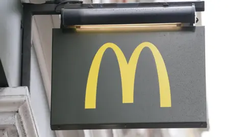 Signage for a branch of McDonald's. It is a dark grey sign with a yellow 'M' and it is it hanging on the wall of a white building.