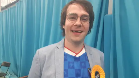 Paul Edgeworth has straight brown hair with a side fringe and blue eyes. He is wearing circular glasses, a blue t-shirt and grey suit jacket with a yellow Liberal Democrat rosette. He is smiling.