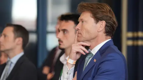 A side view of a man wearing a blue suit with a white shirt and green tie as results are announced at an election count. He has a finger to his mouth and is staring into the distance. There is a union flag badge on his suit lapel.