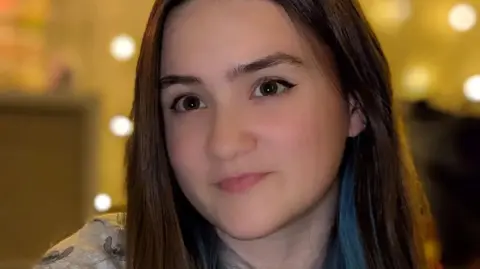 Seren Jaye is pictured with long, straight brown hair with a streak of blue stands indoors against a warm, softly lit background featuring circular light spots. She is wearing a light-coloured top with a subtle pattern. 