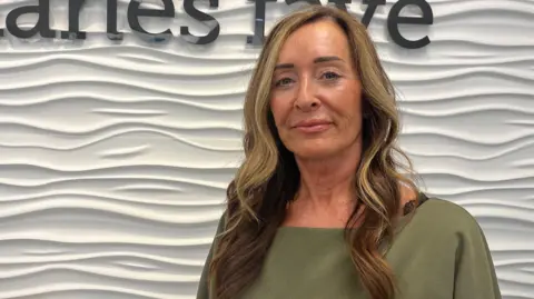 Director of Charles Faye estate agents Louise Burrows with long blonde-brow curled hair and wearing an olive-green blouse standing in front of a white wall.