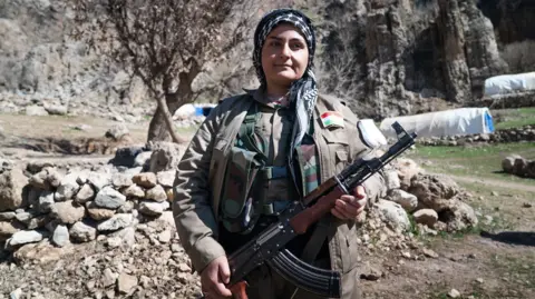 Female Iranian Kurdish fighter dressed in headscarf and grey uniform and holding an automatic weapon stands in front of a pile of stones and some tents in the distance.