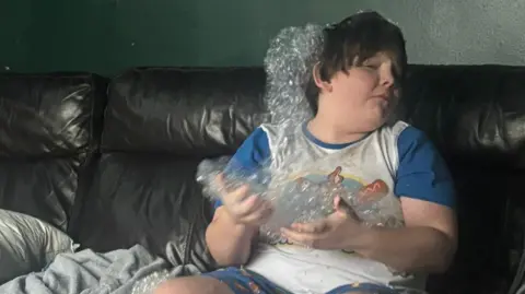 Family handout Rory is playing with bubbles while sitting on a sofa. He is turning his face away from them and looks anxious. He is wearing short-sleeved pyjamas that are grey and blue.